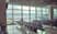 Large airport waiting area with rows of empty black seats facing tall floor‑to‑ceiling windows, where part of an airplane is visible outside on the tarmac.